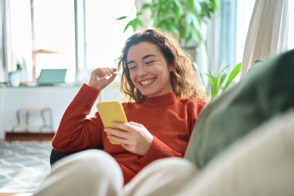 A young woman with curly hair smiles while sitting comfortably on a couch, holding a yellow smartphone. She appears relaxed and engaged, with natural light illuminating the cozy indoor setting adorned with greenery.