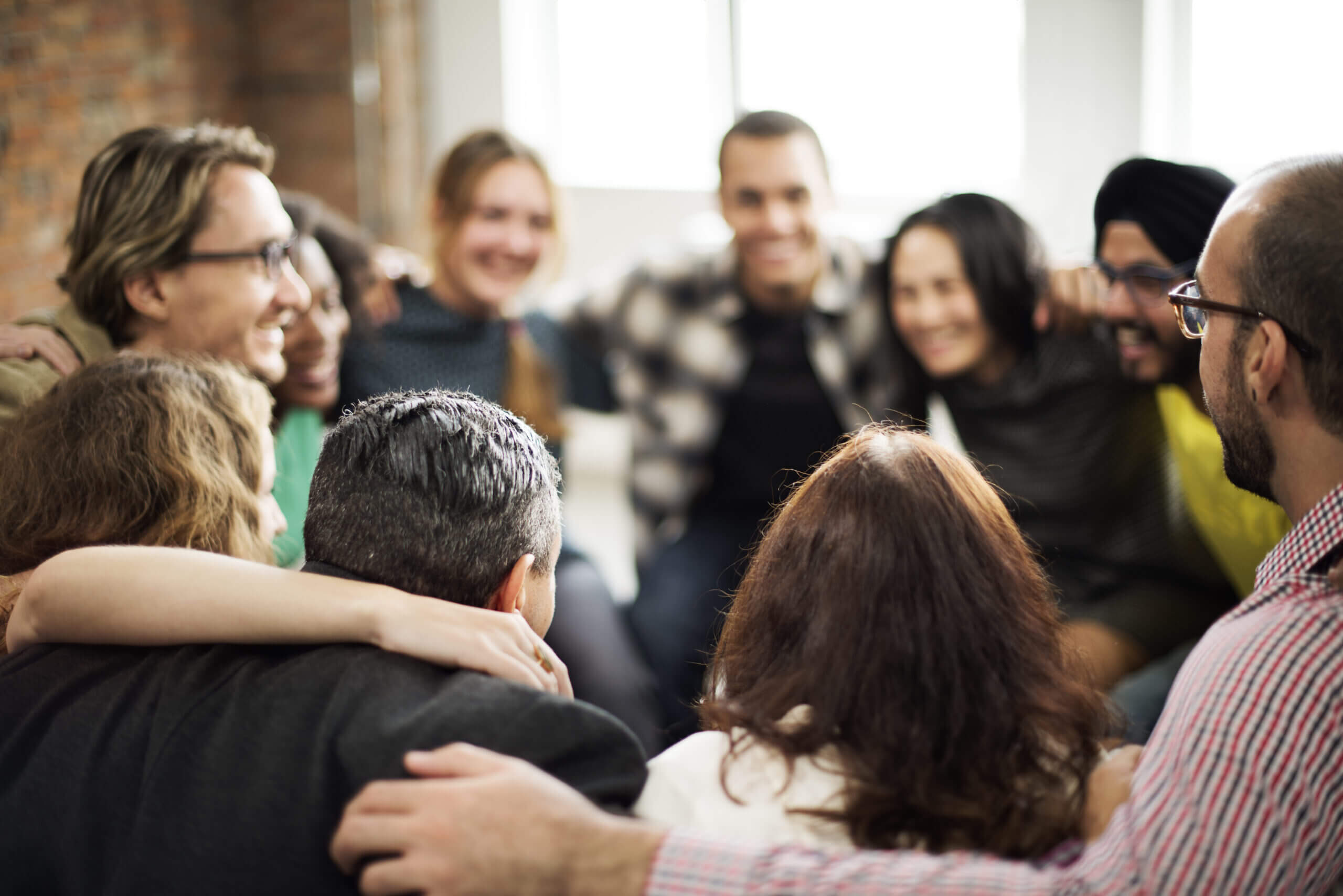 A diverse group of people are gathered in a circle, smiling and engaging in a warm, friendly discussion. The setting appears relaxed and inviting, with a mix of casual attire and visible camaraderie among the participants.
