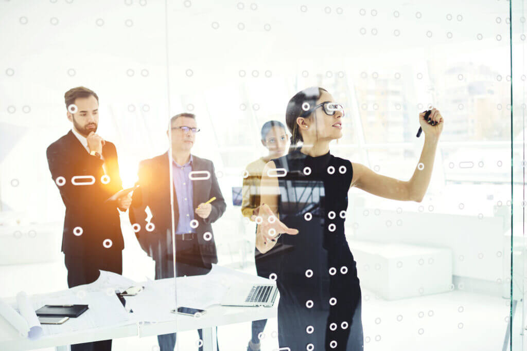 A woman in a black shirt presents ideas on a glass wall while three men in business attire observe. The scene is bright and modern, featuring a collaborative work environment.