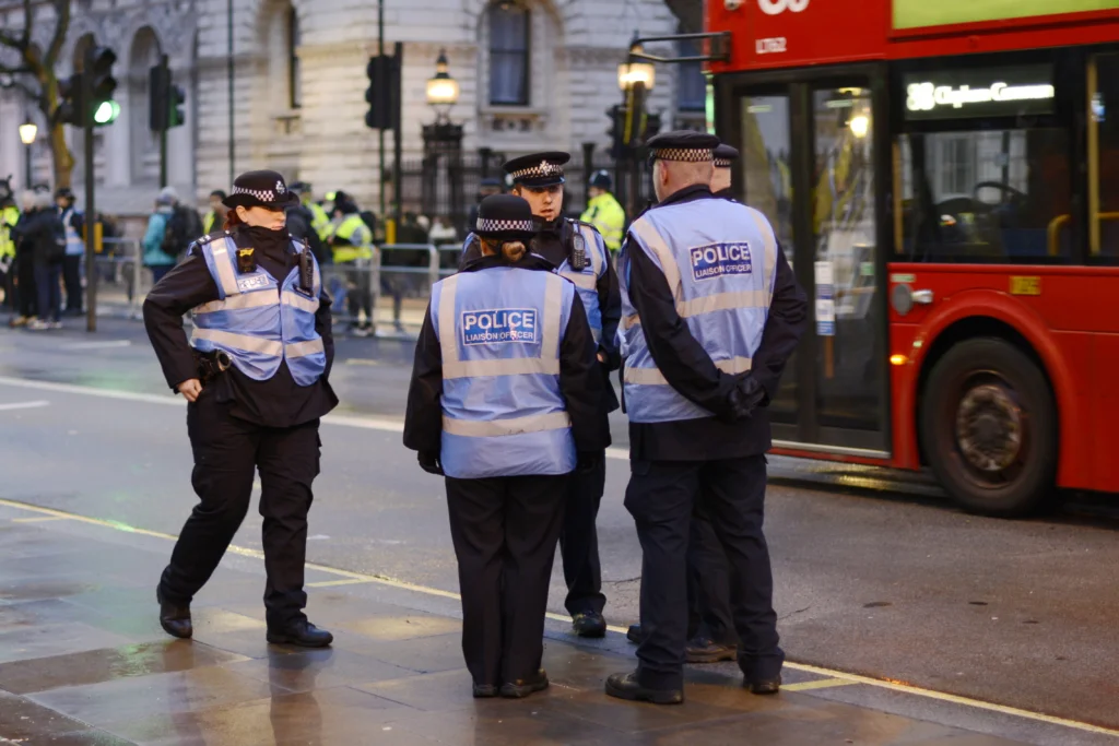 Police officers in reflective vests stand on a city street near a red double-decker bus.