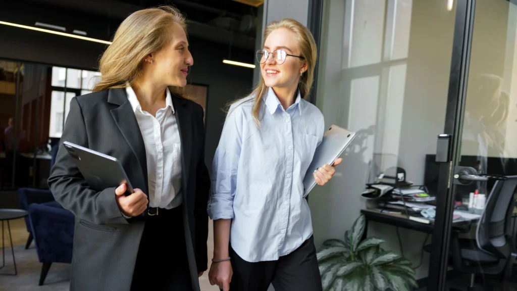 Two women, one in a suit and the other in a blouse, walk through an office while holding tablets and smiling at each other.